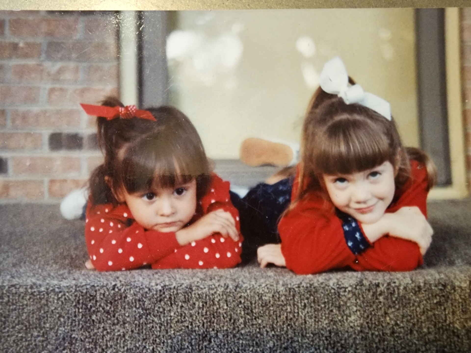 Nikki and Ali as toddlers lying side by side in matching red polka-dot dresses with white bows.