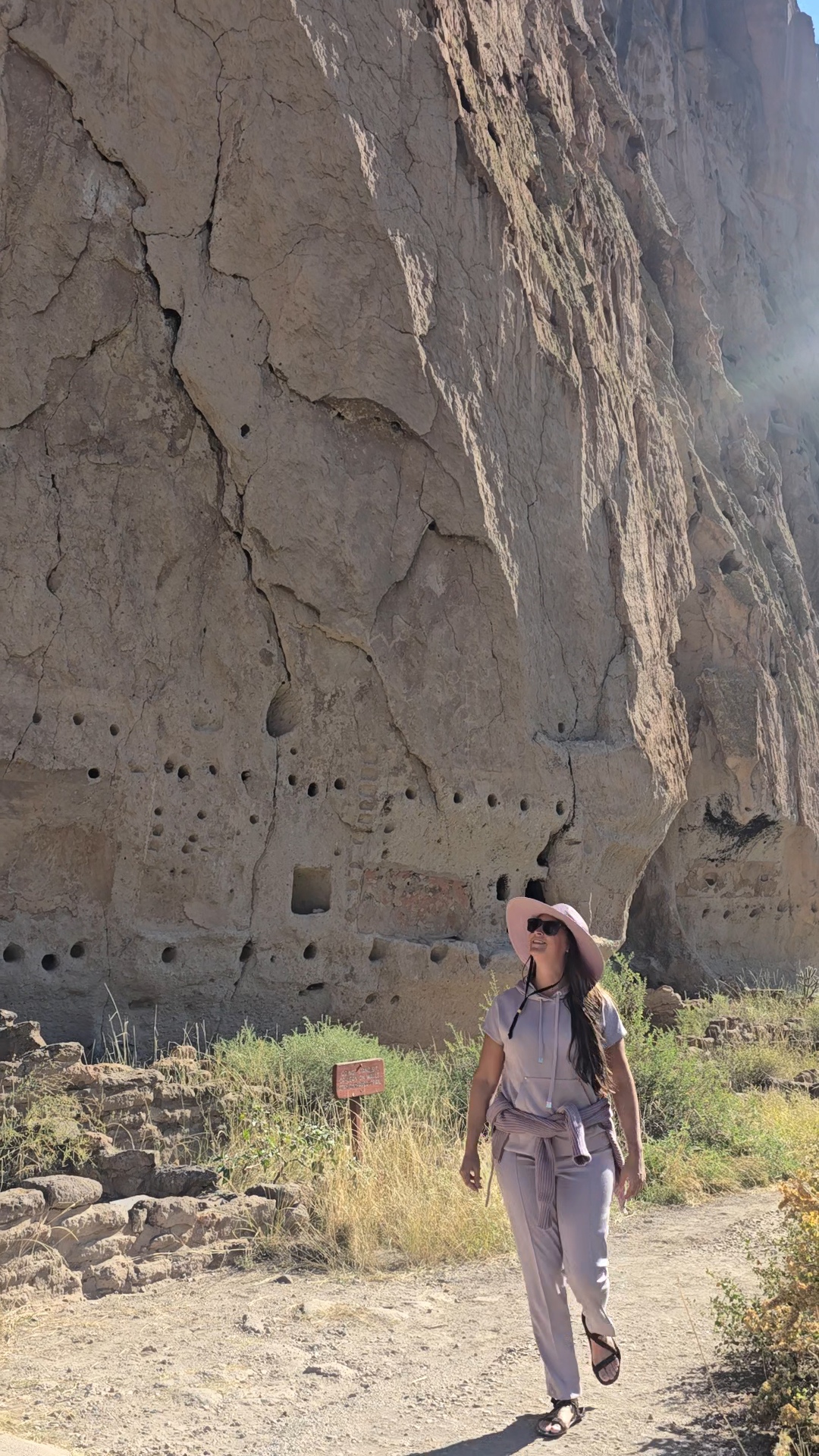 Nikki Bodie in a wide-brimmed hat walking beneath the carved cliff dwellings at Bandelier.