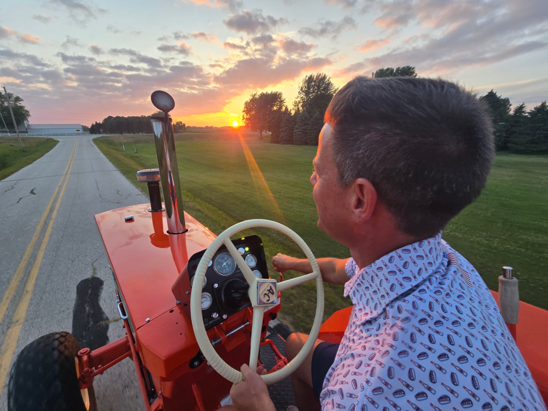 A man driving a bright orange Allis-Chalmers tractor down a Wisconsin country road at sunset.