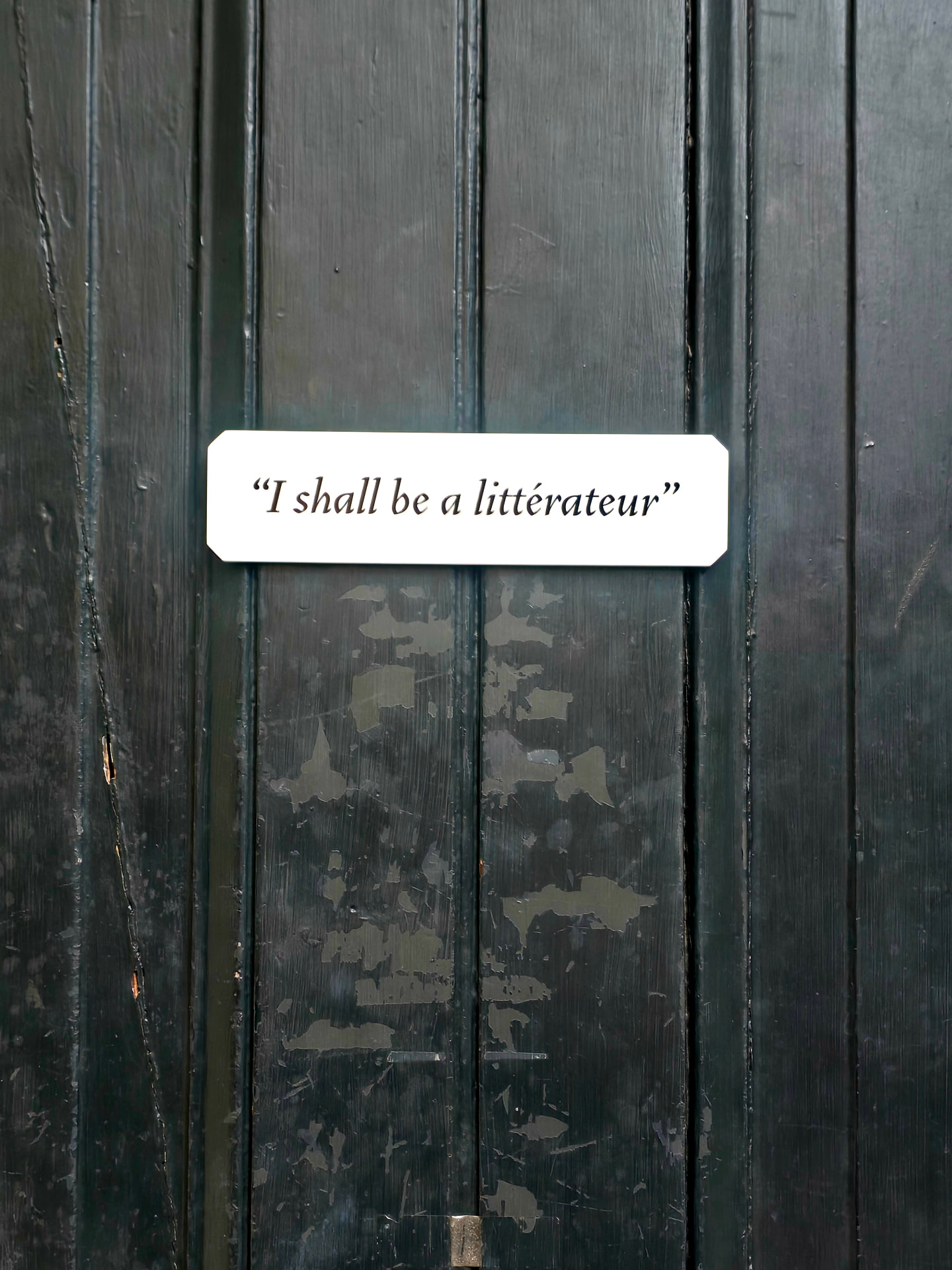 A weathered dark green wooden door with a small white plaque reading "I shall be a littérateur."
