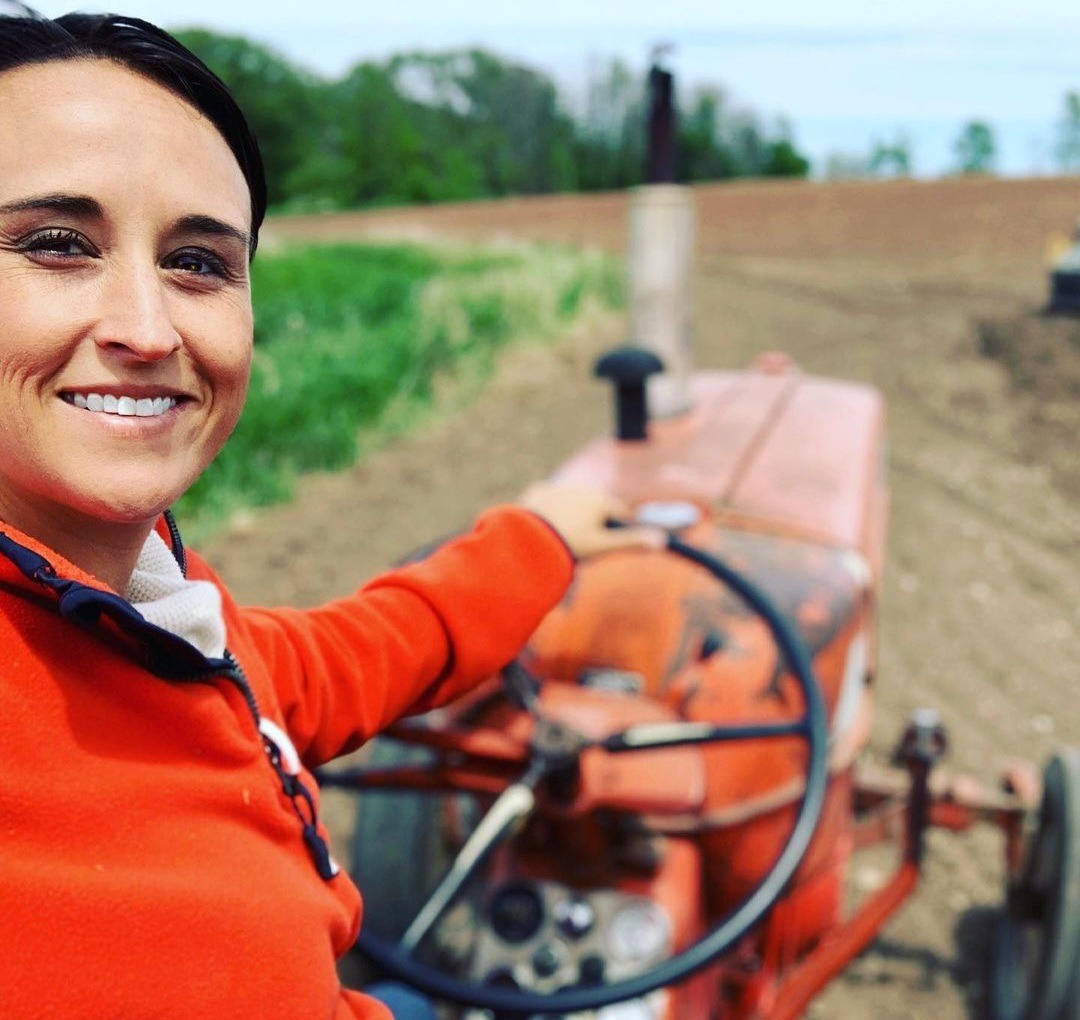 Nikki Bodie smiling from the seat of an old orange tractor in a plowed field.