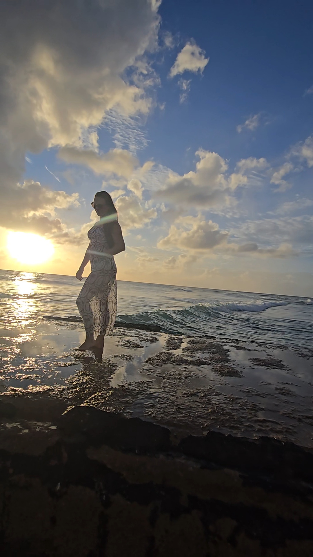 Nikki Bodie standing in shallow water on the reef rock at sunset, clouds and blue sky above.