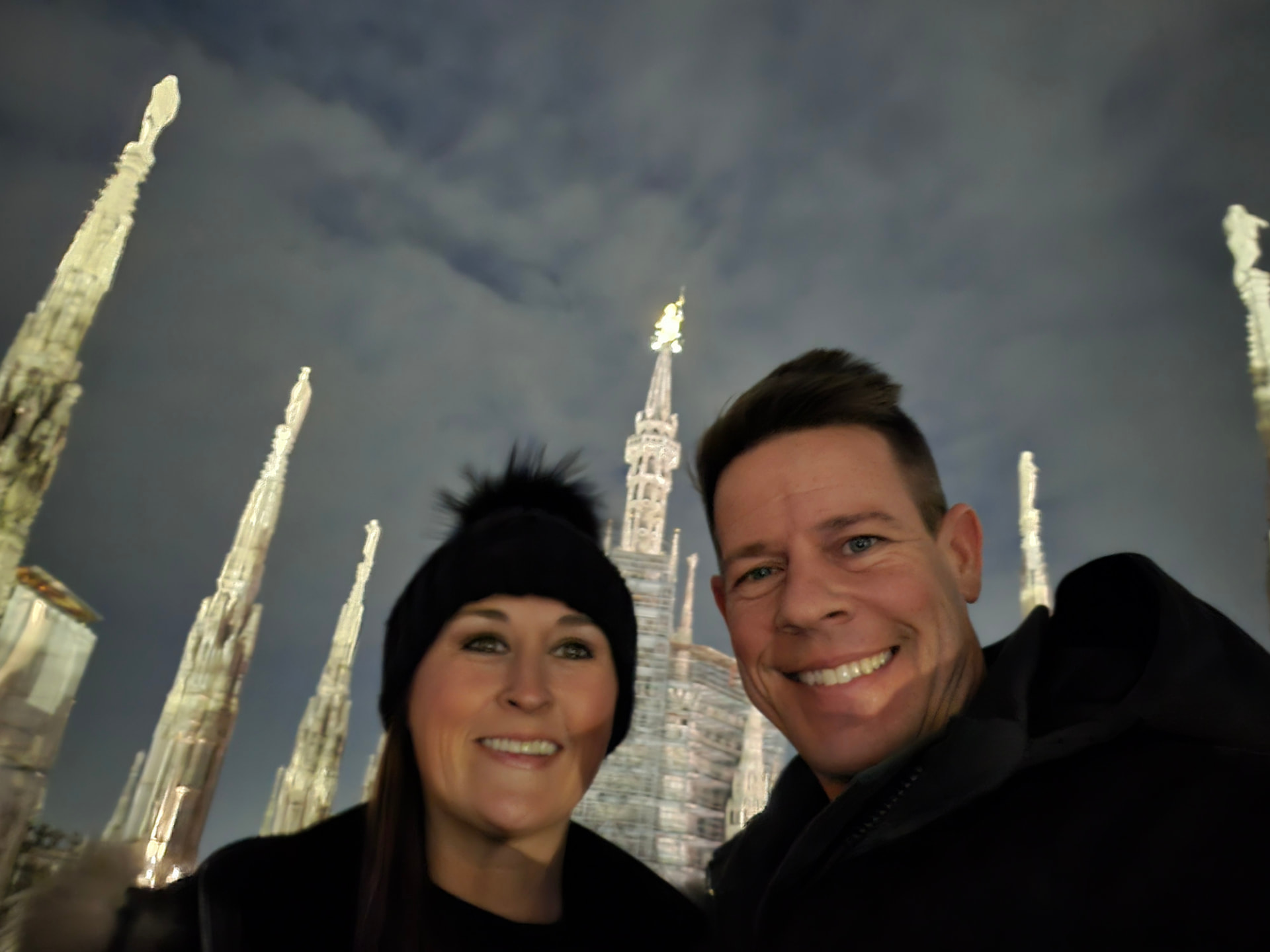 Nikki Bodie and Quentin smiling in a selfie among the lit spires of the Duomo rooftop at night.