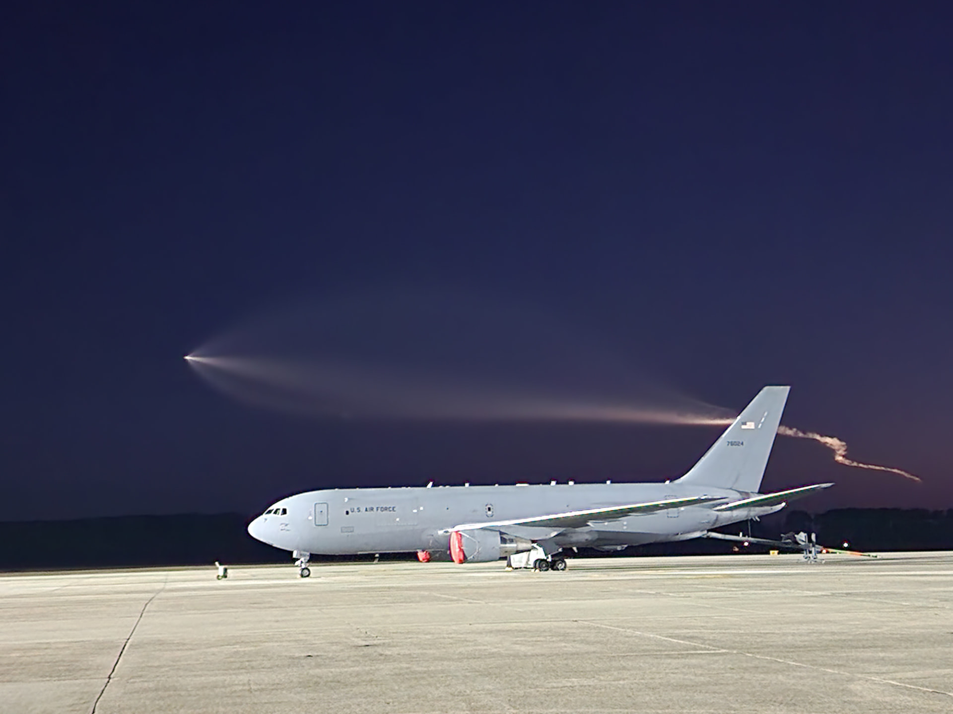 A KC-46 parked at night as a rocket trail streaks across the sky.
