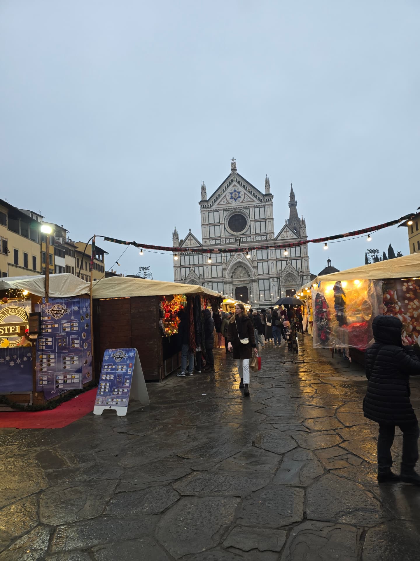 The Florence Christmas market on wet stone in the rain, Santa Croce rising behind the stalls.