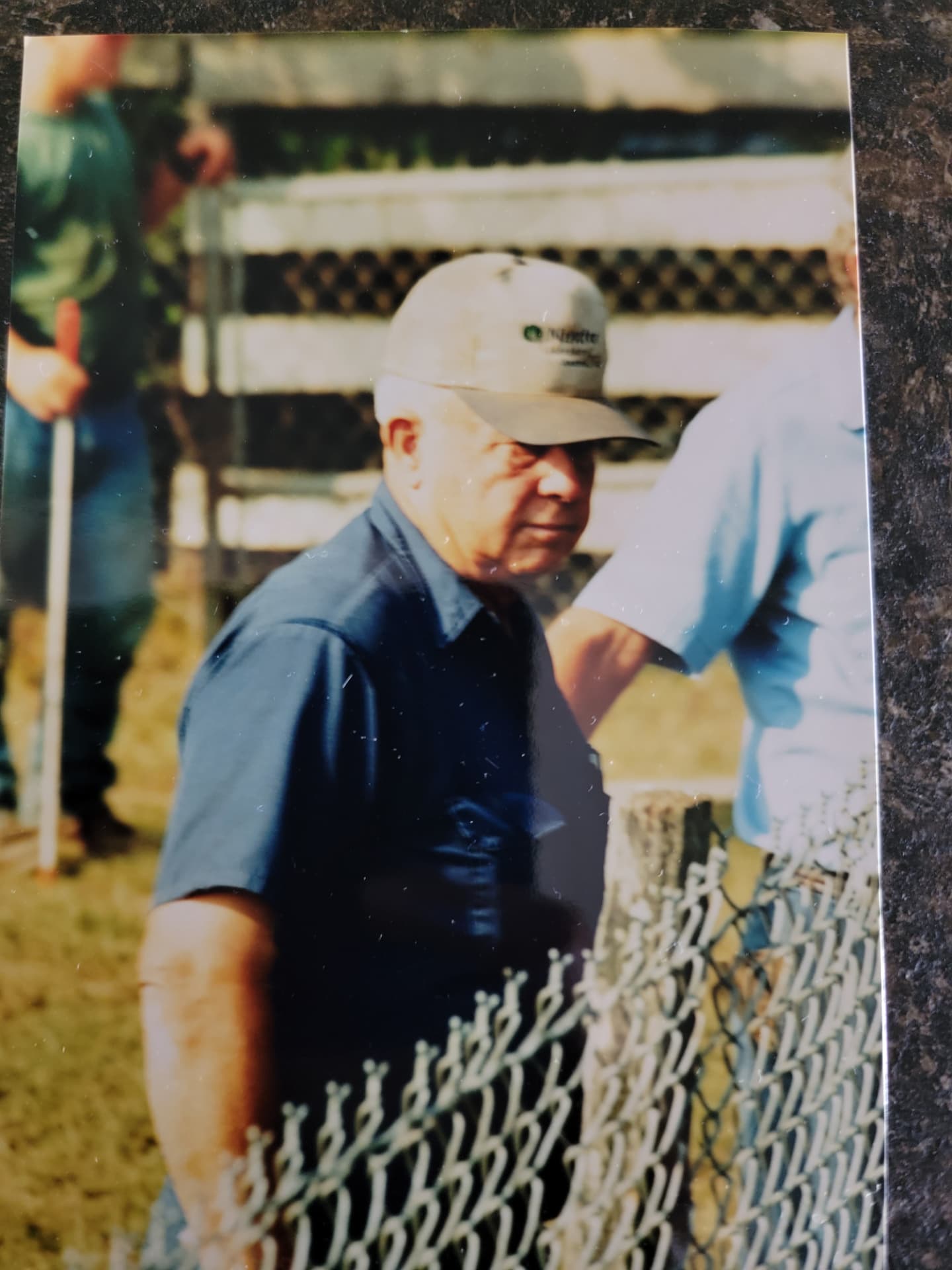 A faded color photograph of an older man in a cap and blue work shirt at a chain-link fence on the family farm.