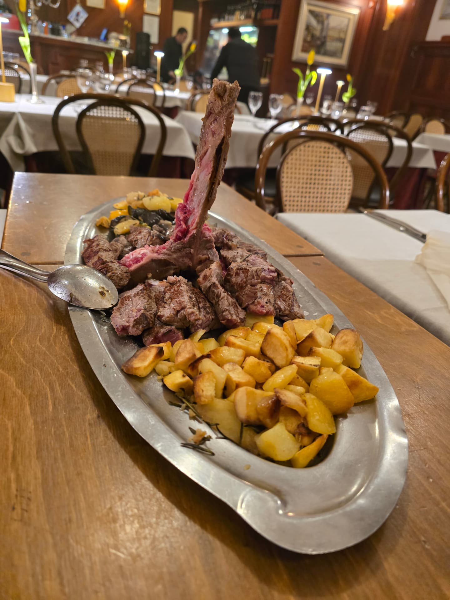 A Florentine steak on the bone with roast potatoes on a metal platter in a traditional restaurant.