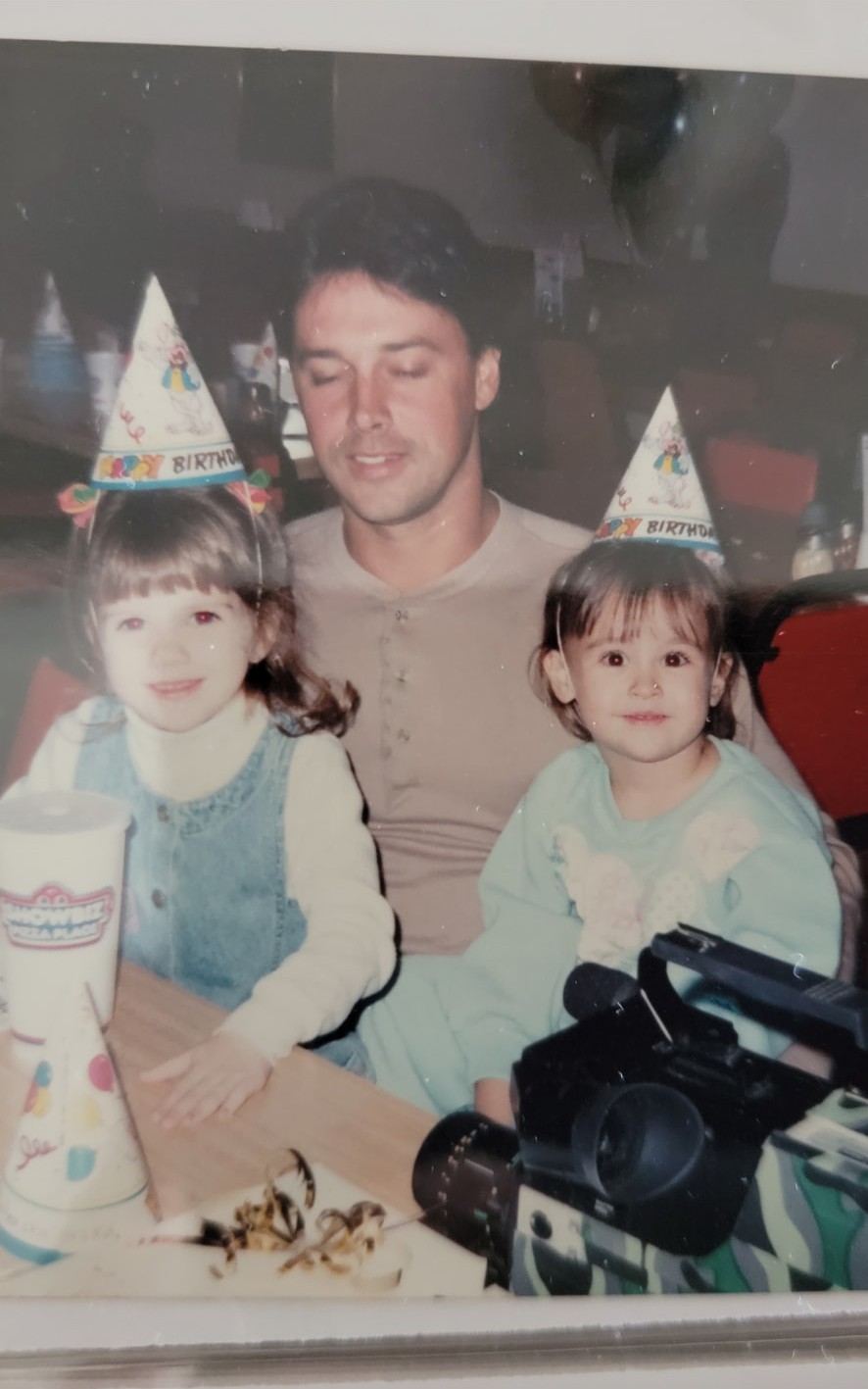 Nikki and Ali as toddlers in party hats at a birthday table with a young father.