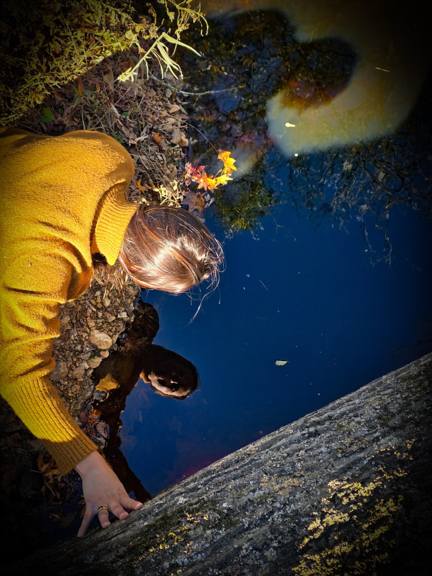 Seen from above, Nikki Bodie in a mustard sweater leans over dark creek water, her reflection looking back up at her.