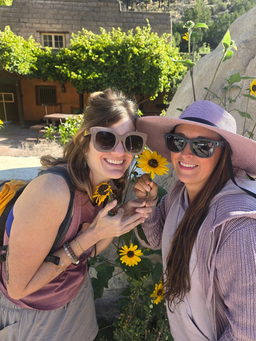 Nikki and Ali grinning cheek to cheek beside tall sunflowers in New Mexico.