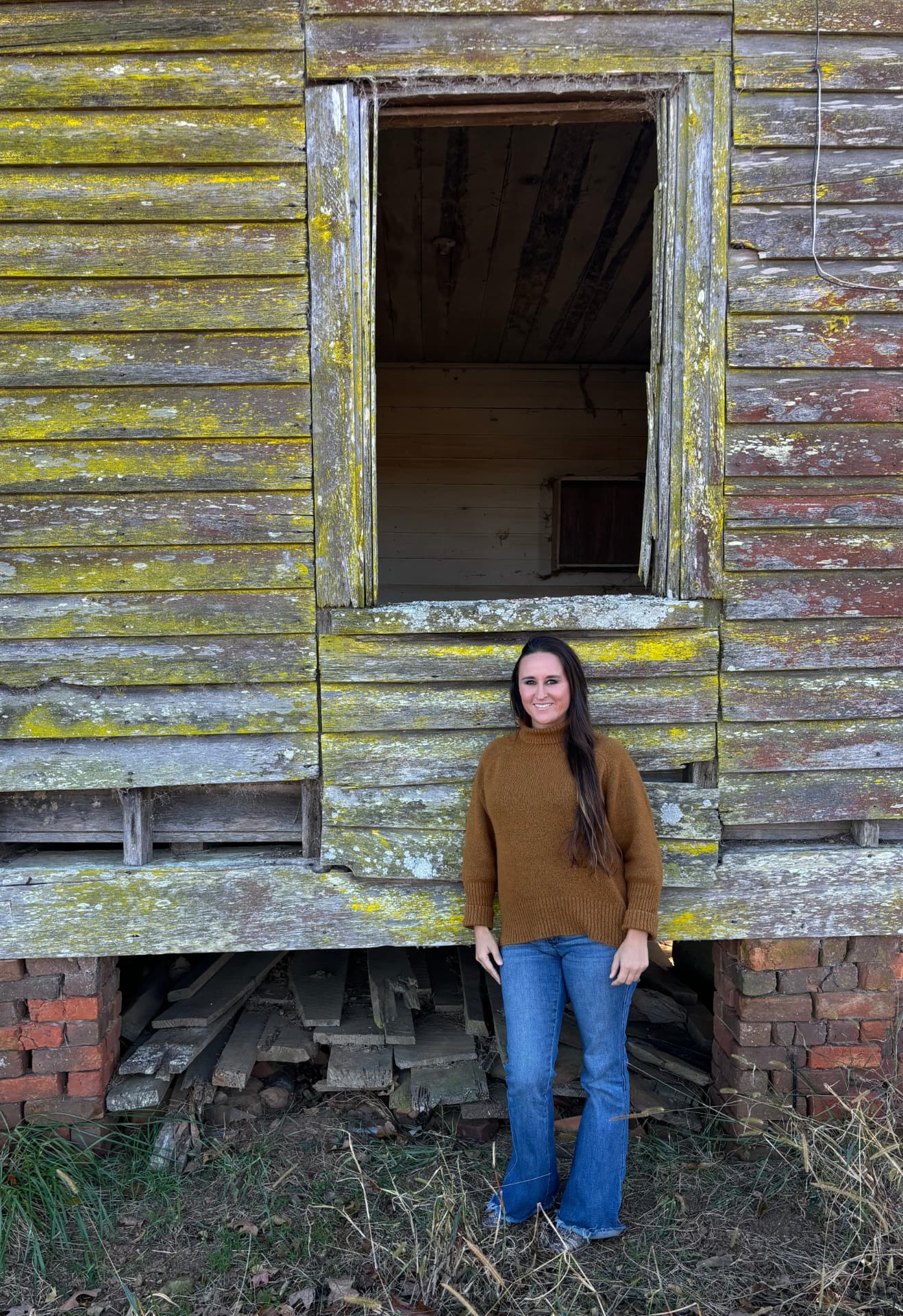 Nikki Bodie standing in front of an old weathered wooden building covered in lichen, beneath a large empty window opening.