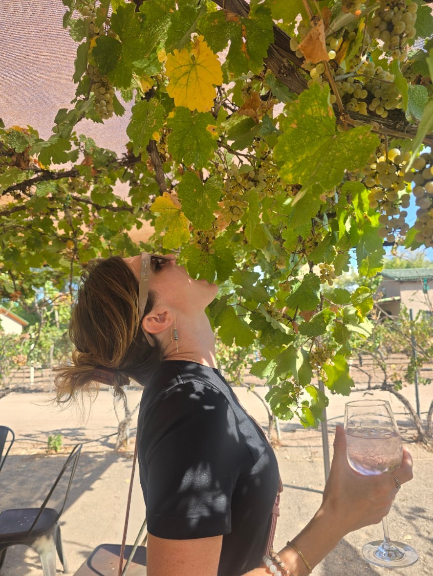 Ali tipping her head back to smell a cluster of grapes hanging from the vines.