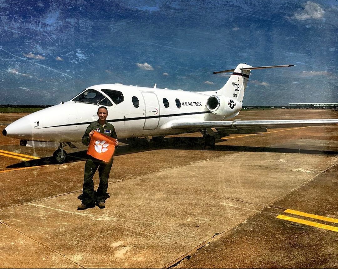 Nikki in her flight suit holding a Clemson tiger-paw flag beside a T-1 jet.