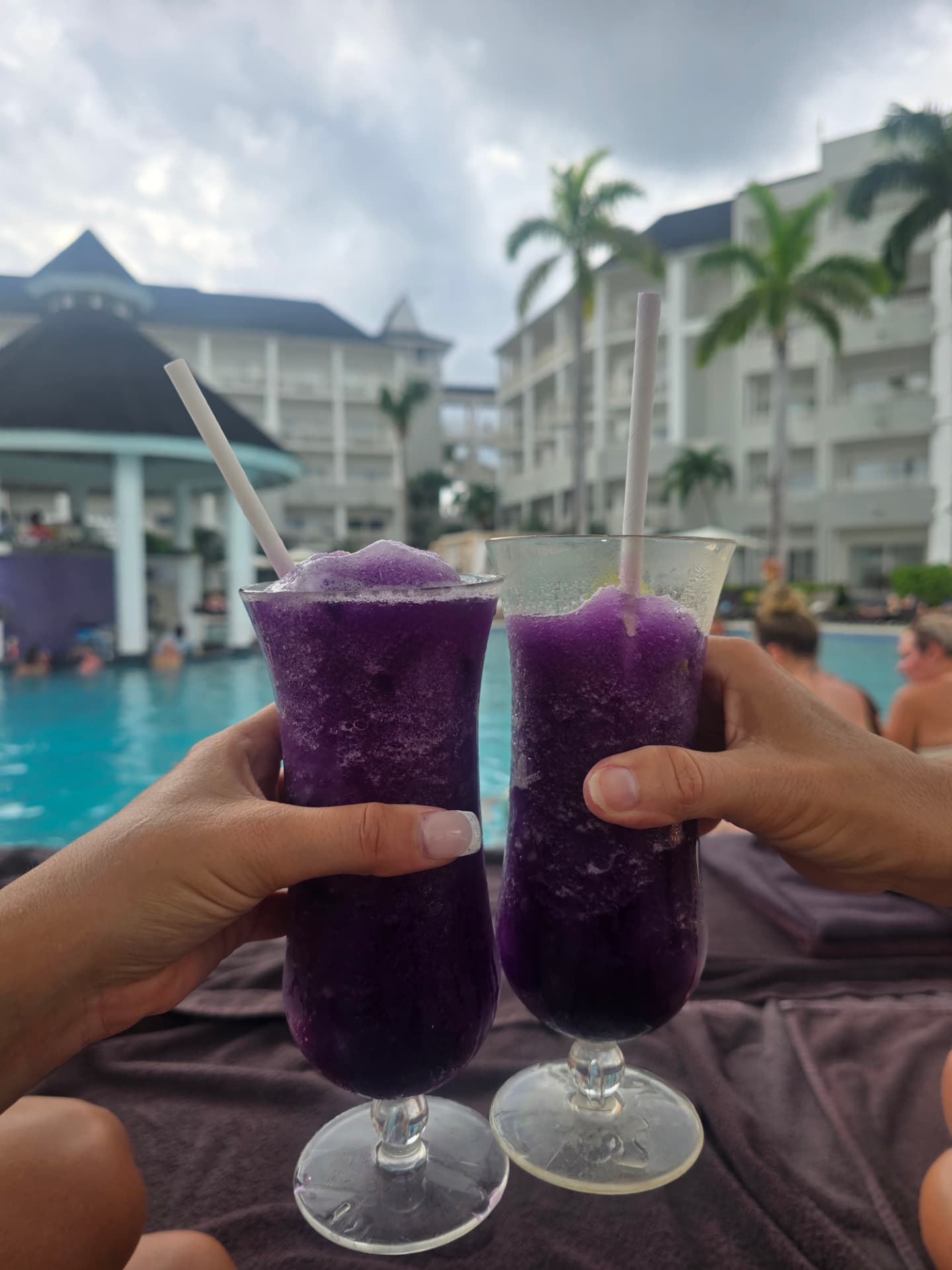Two hands touching glasses of purple frozen drinks beside the resort pool.