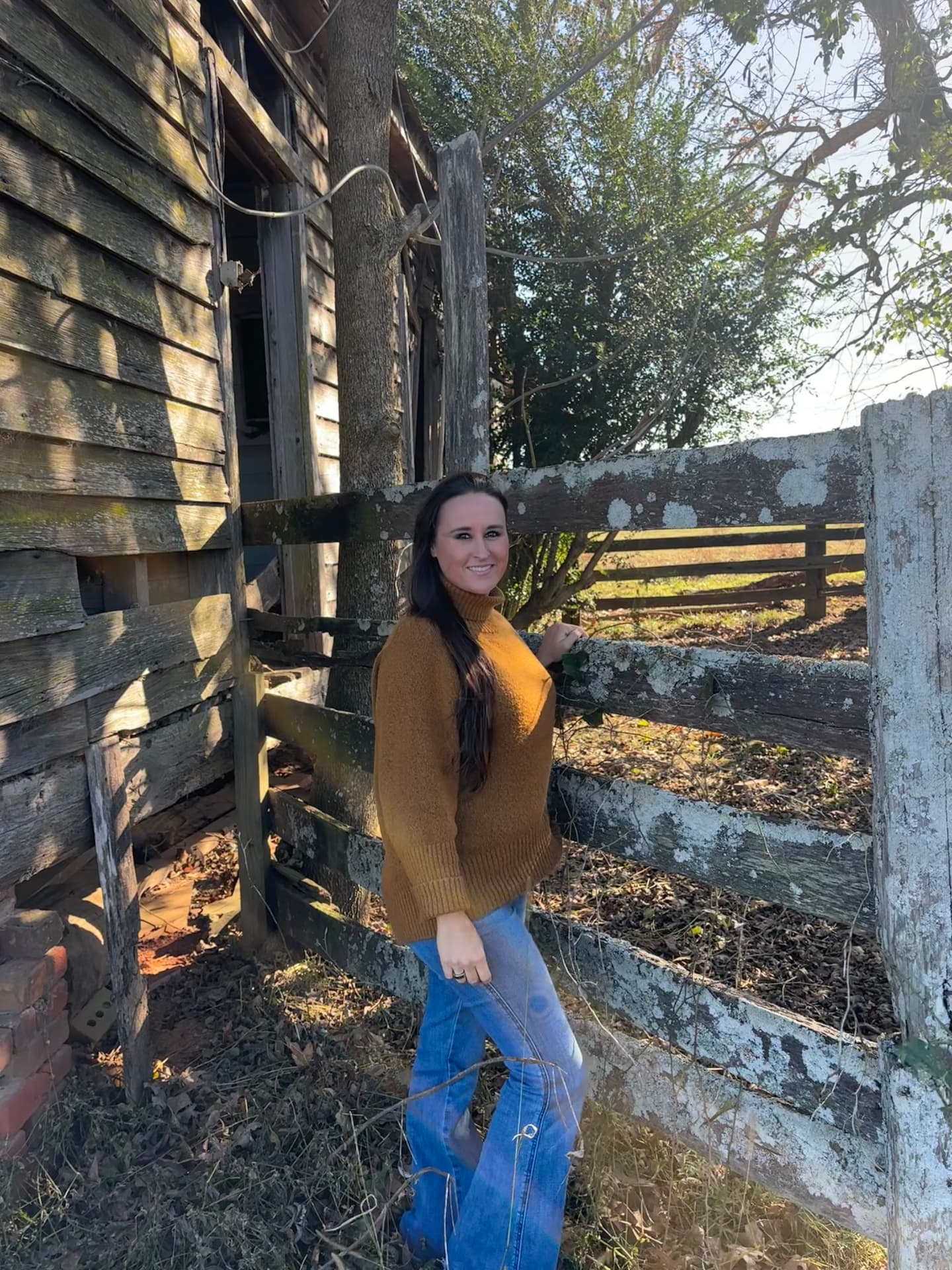 Nikki Bodie leaning on an old lichen-covered wooden fence beside the weathered building in fall sun.