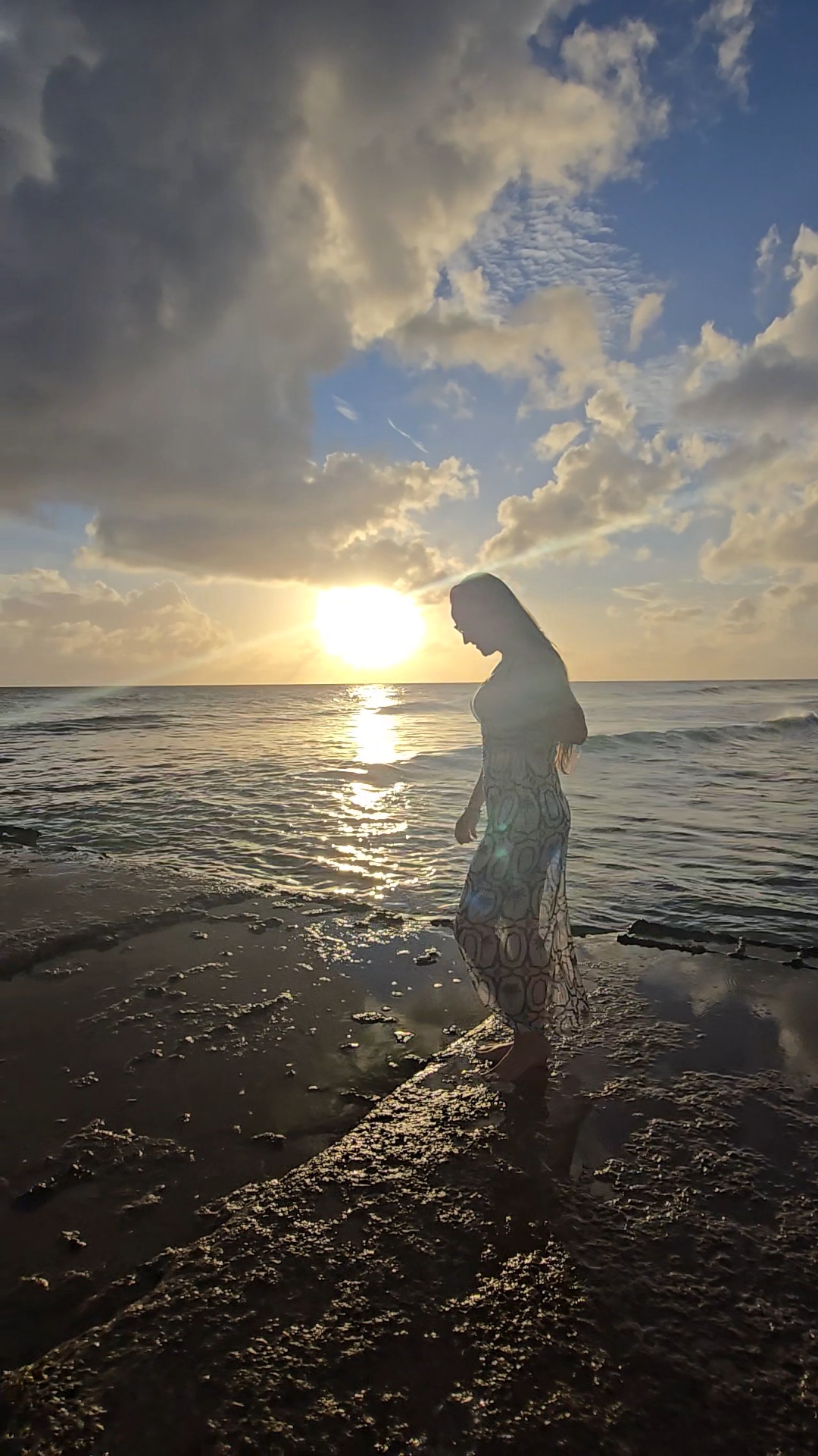 Nikki Bodie in silhouette at the water's edge, looking down, with the sun setting behind her over the sea.