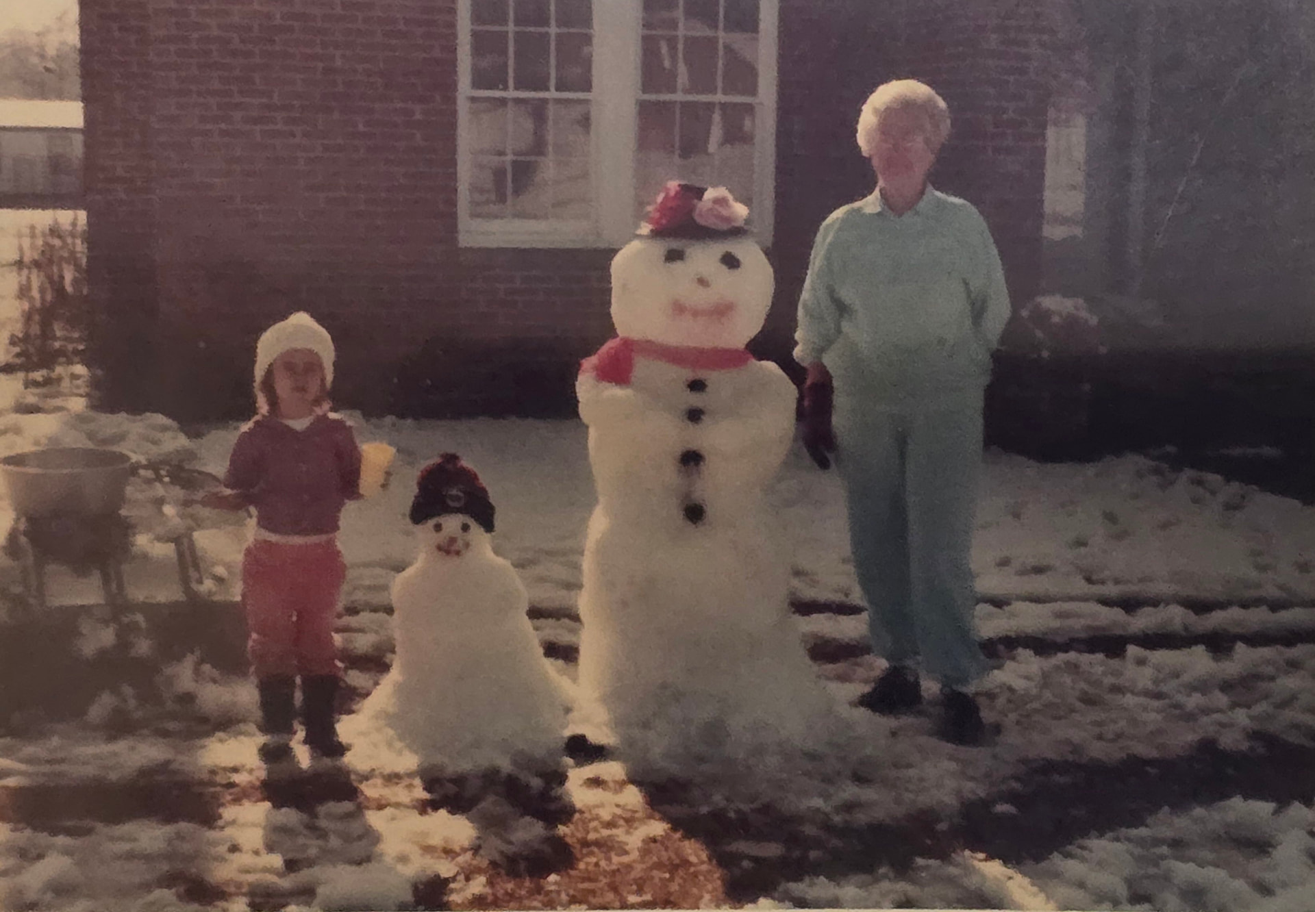 A faded color photograph of young Nikki and Grandma Reba beside two snowmen they built in front of a brick house.