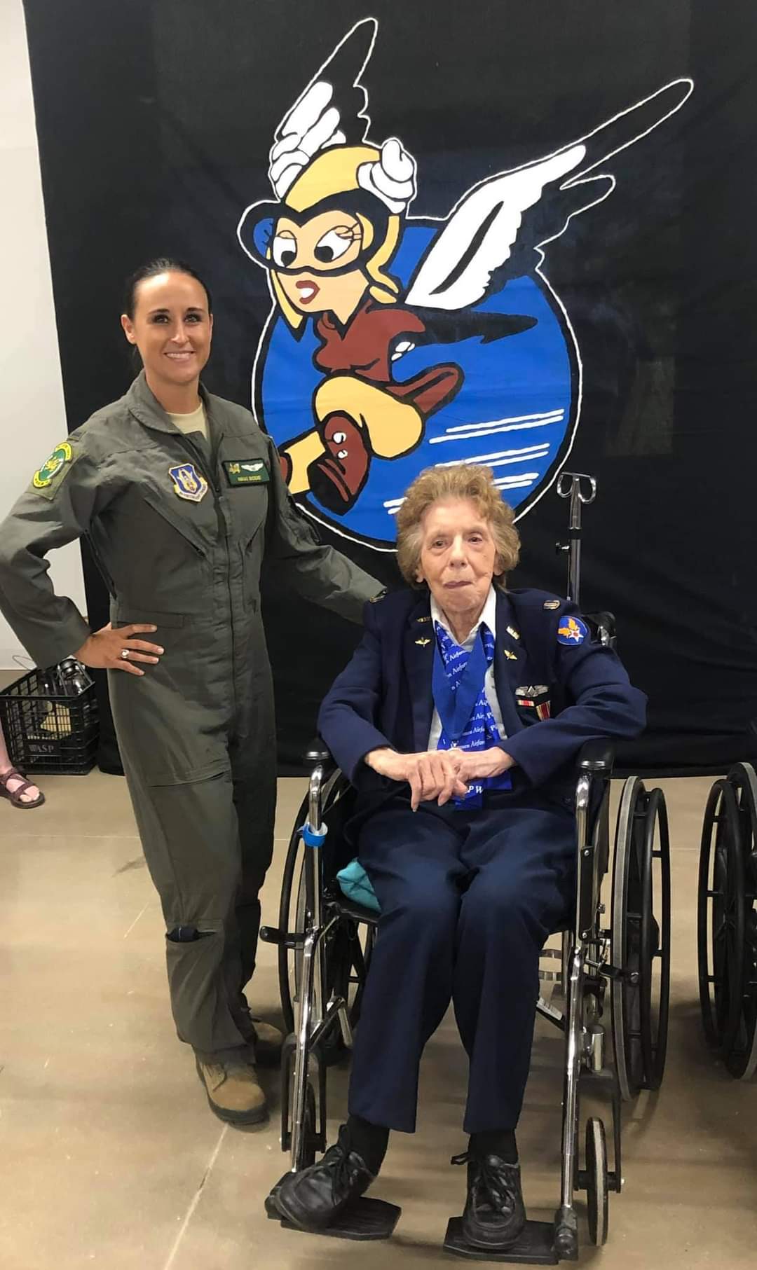 Nikki in her flight suit beside a WASP in uniform, in front of the winged WASP mascot banner.