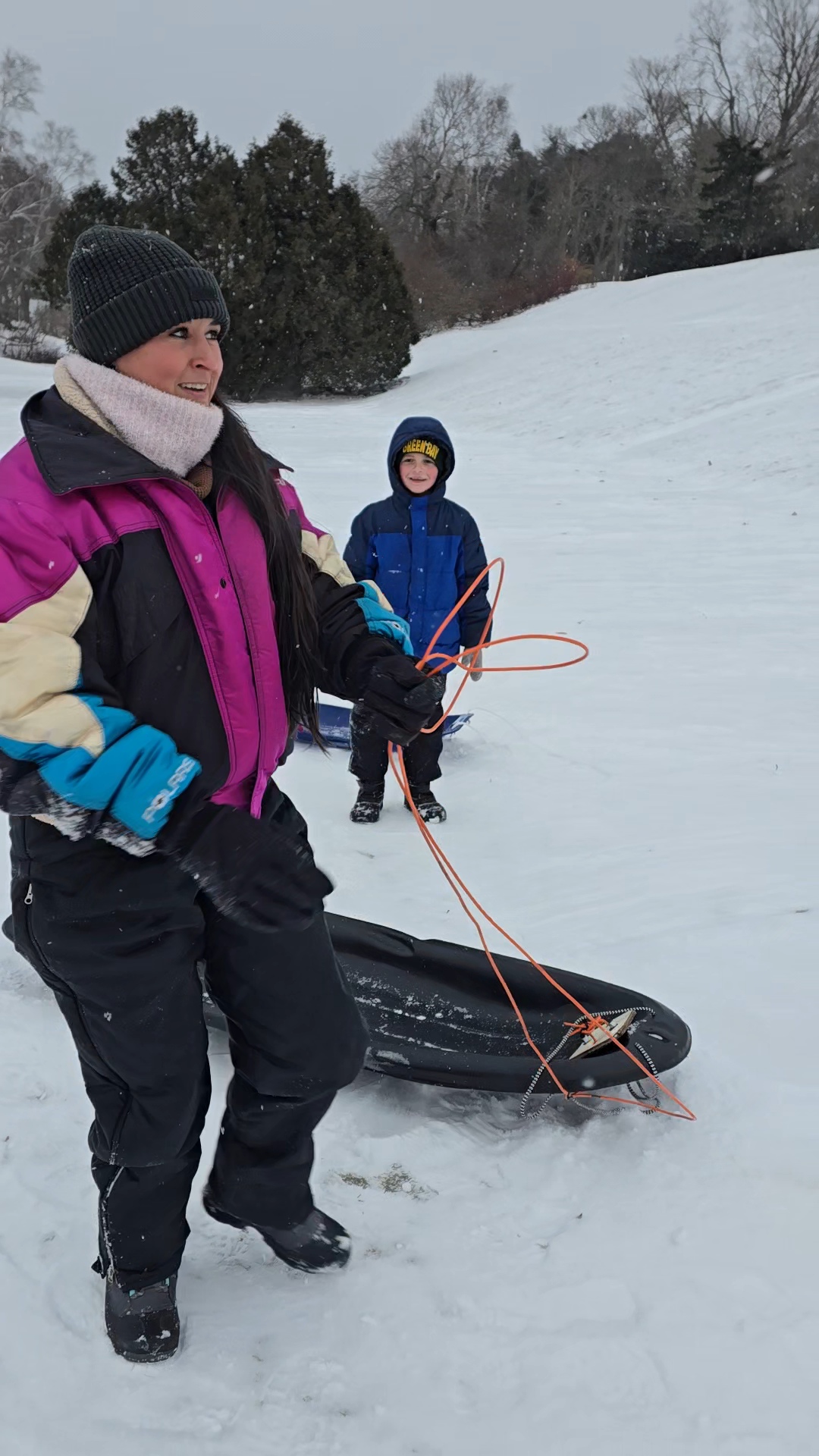Nikki Bodie holding an orange sled rope on a snowy hill, a boy in a Green Bay hat smiling behind her.