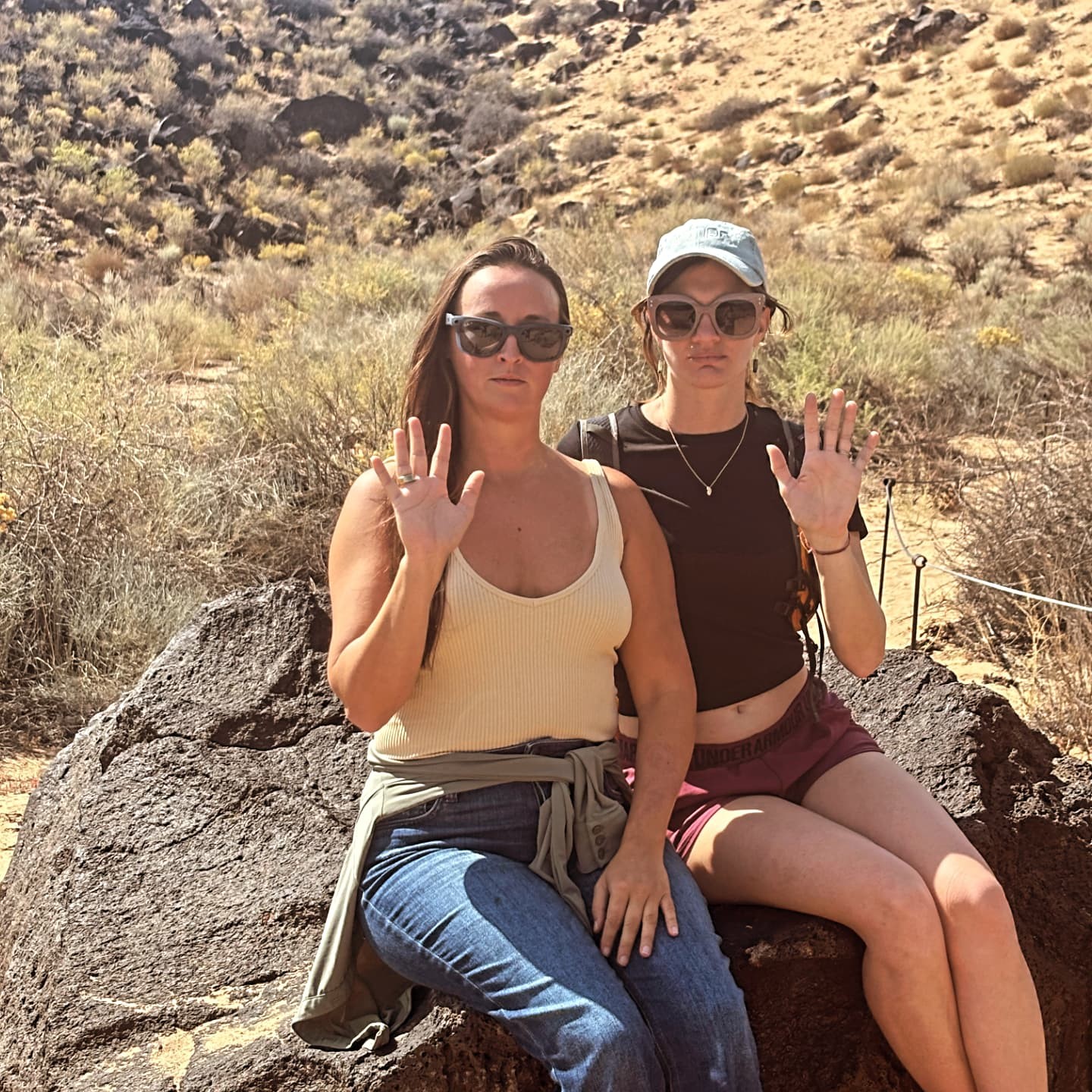 Nikki and Ali sitting on a desert boulder, each with one hand raised.