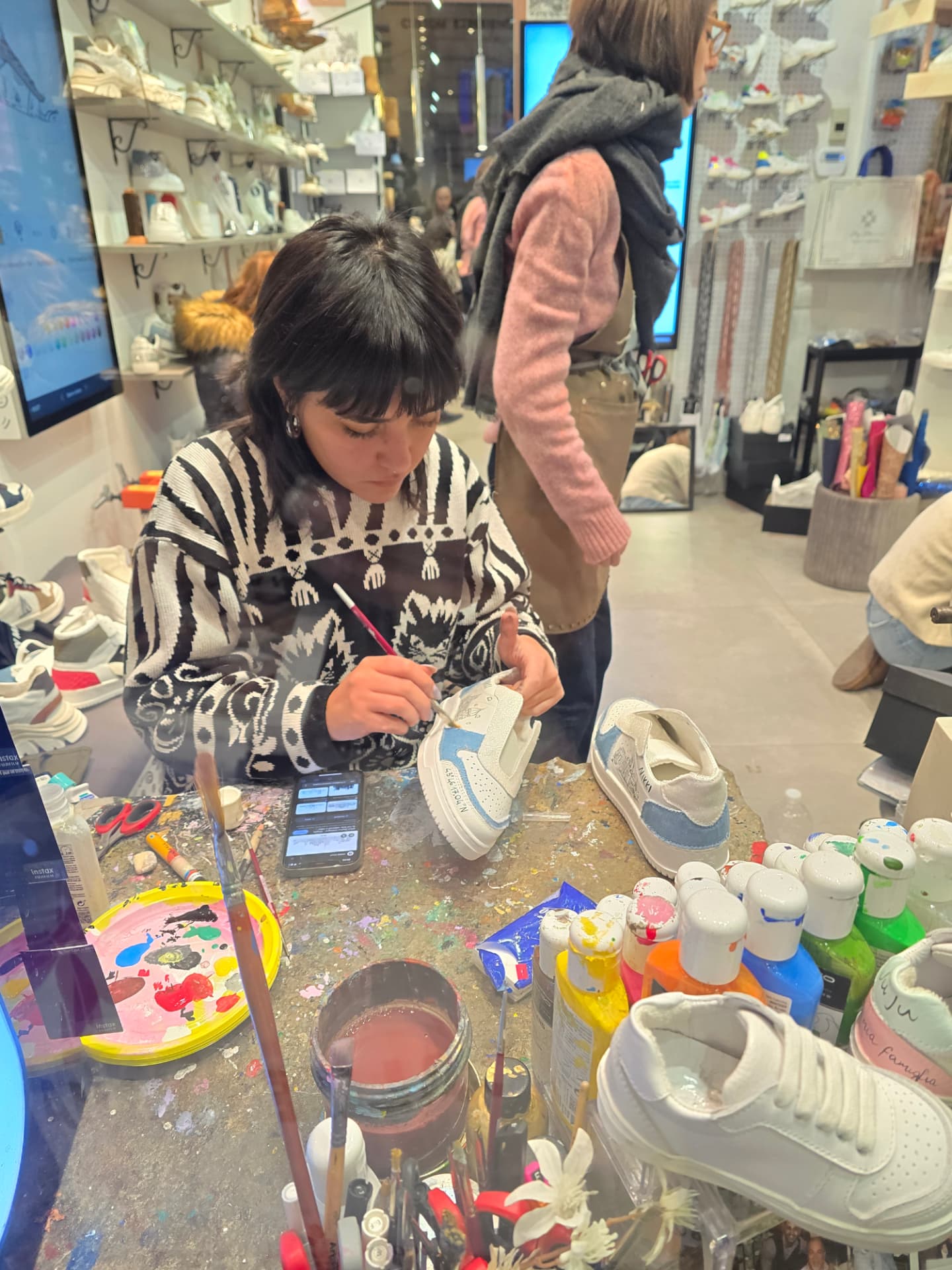 An artist hand-painting white sneakers at a crowded worktable of paints and brushes.