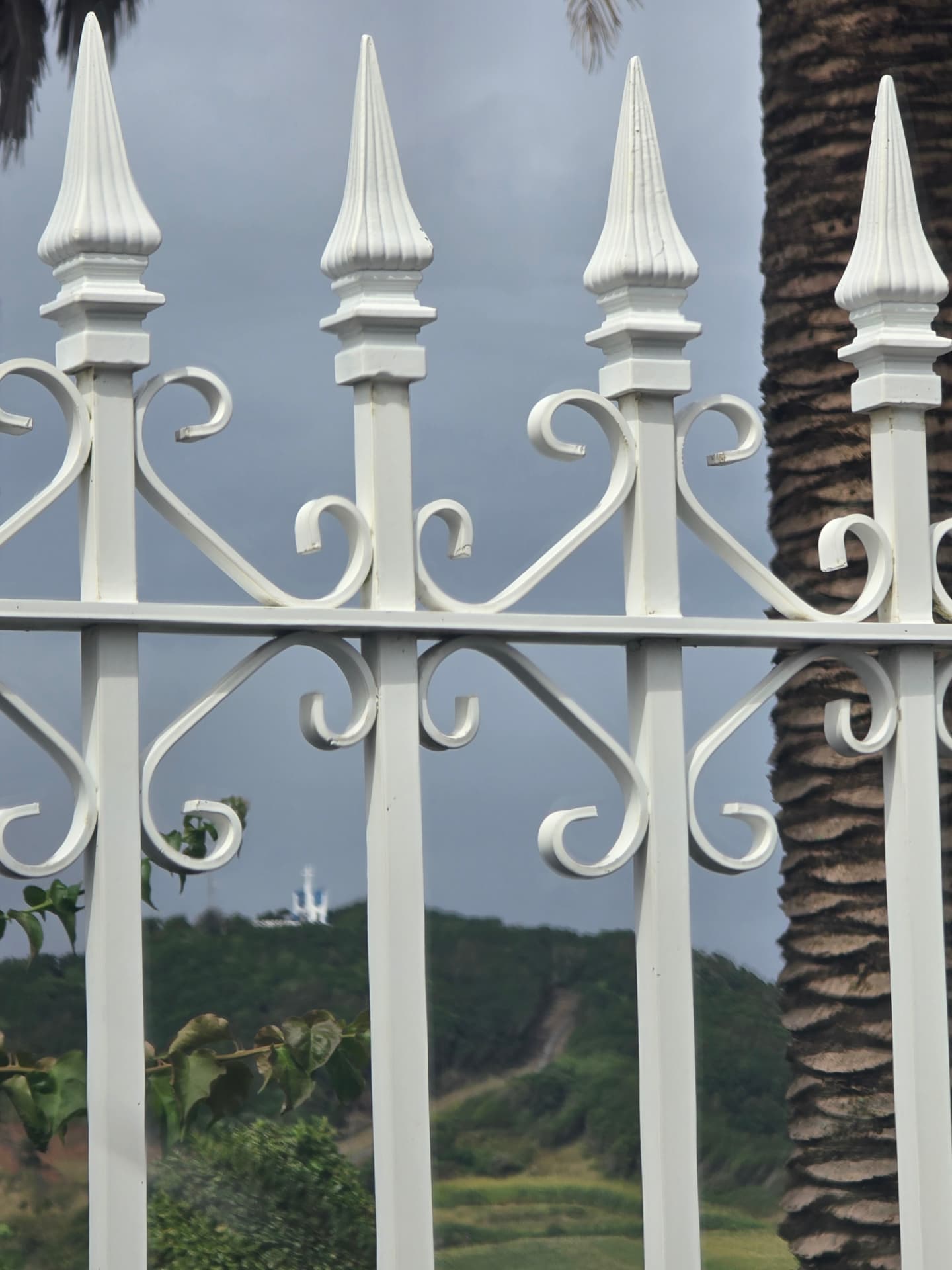 White iron fence finials with a green hill beyond.