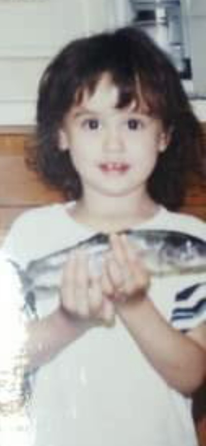 Nikki at three holding up her first fish with both hands — the same fish Grandma Reba kept for decades and that now rests in her Mason jar.
