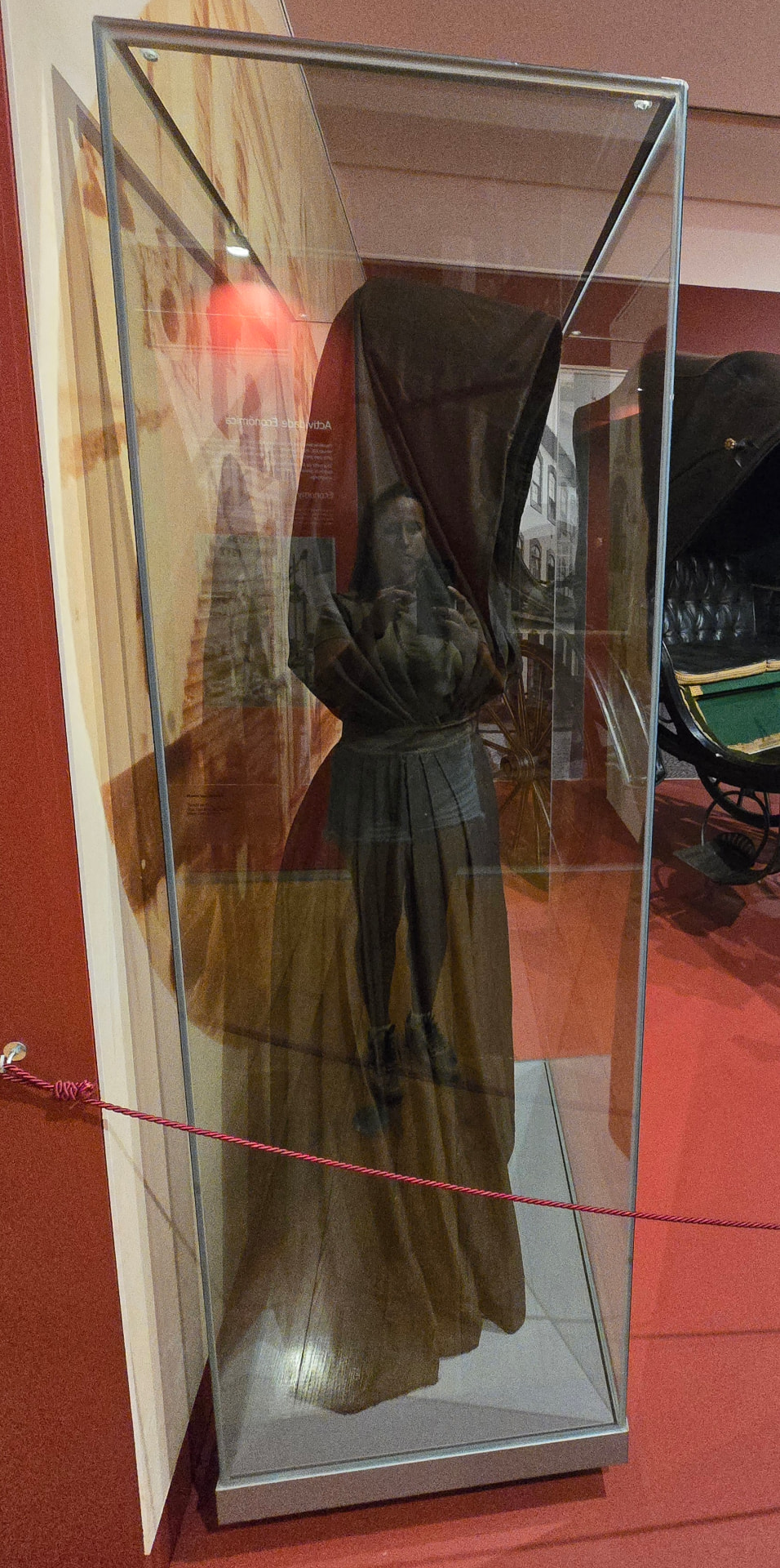 An original capote e capelo displayed upright in a glass museum case, a figure reflected in the glass.