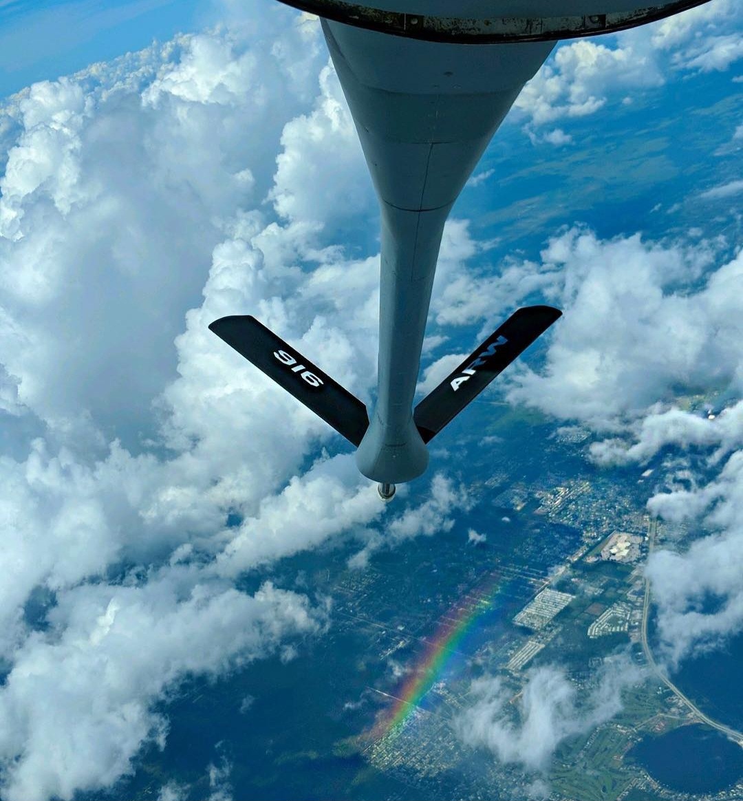 The refueling boom above clouds with a rainbow on the ground far below.