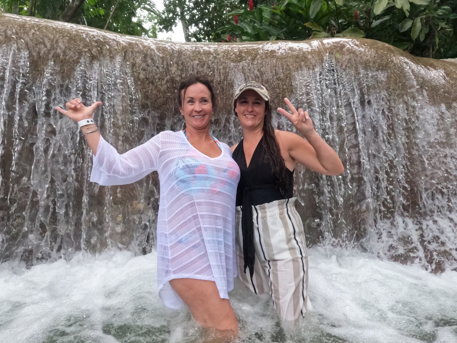 Patti and Nikki in the rushing waterfall, Patti grinning with arms up, Nikki flashing a peace sign.
