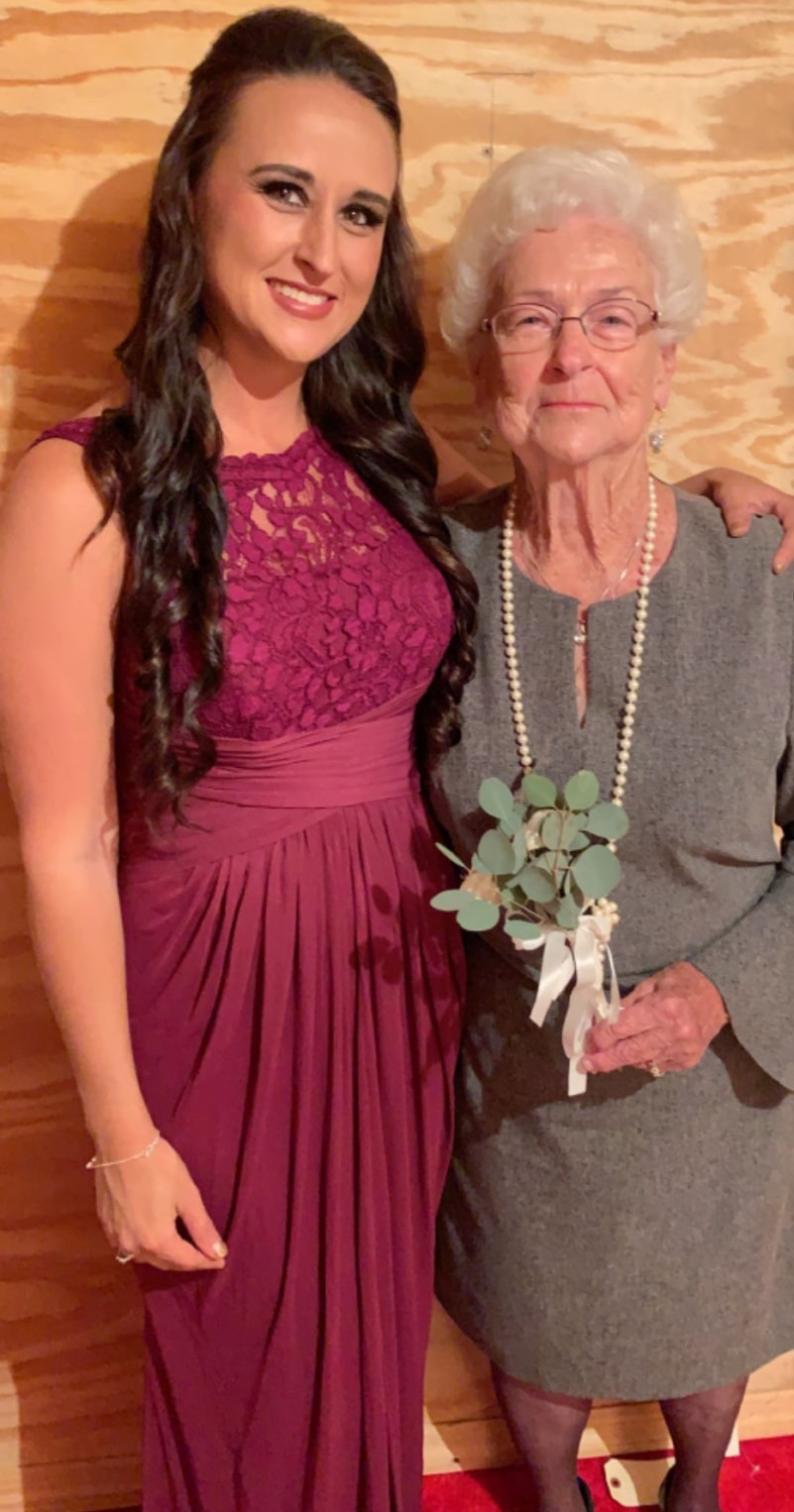 Nikki in a burgundy dress standing beside Grandma Reba in gray, holding a small eucalyptus bouquet.