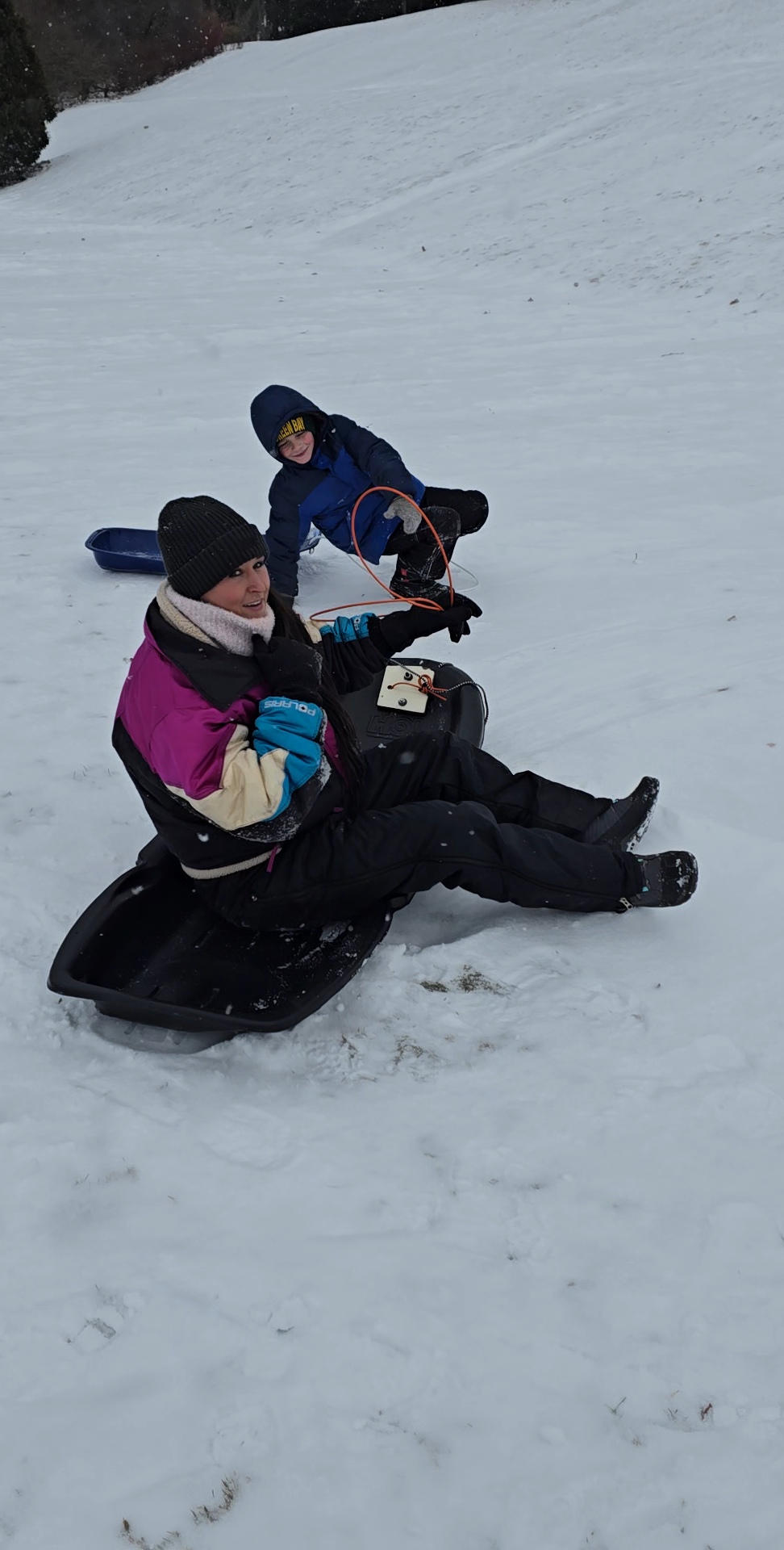 Nikki Bodie sitting on a black sled in the snow, looking back, with a boy on the hill behind her.