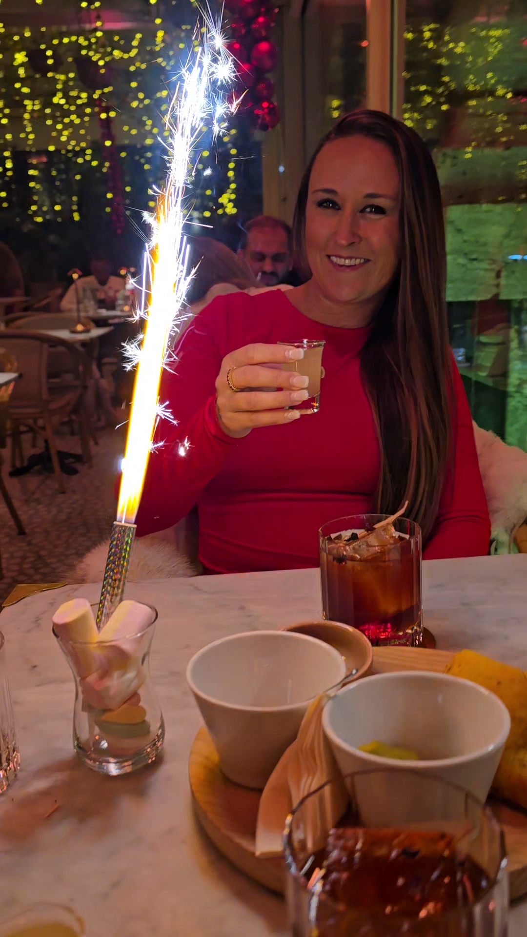 Nikki Bodie in red smiling behind a birthday sparkler, holding up a drink at a restaurant table.
