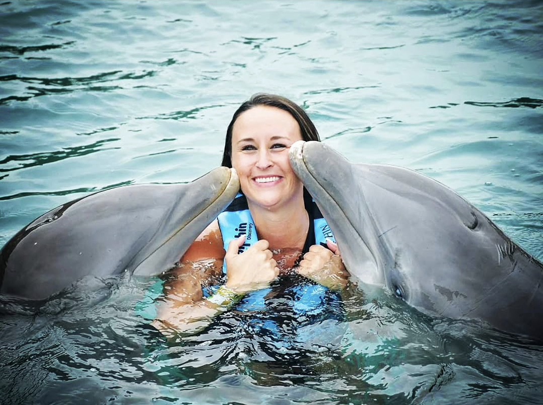 Nikki smiling in the water as two dolphins kiss her on each cheek.