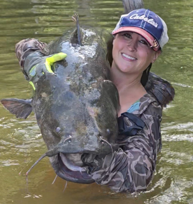 Nikki Bodie in a camouflage jacket and cap grinning as she lifts a huge catfish out of the water.