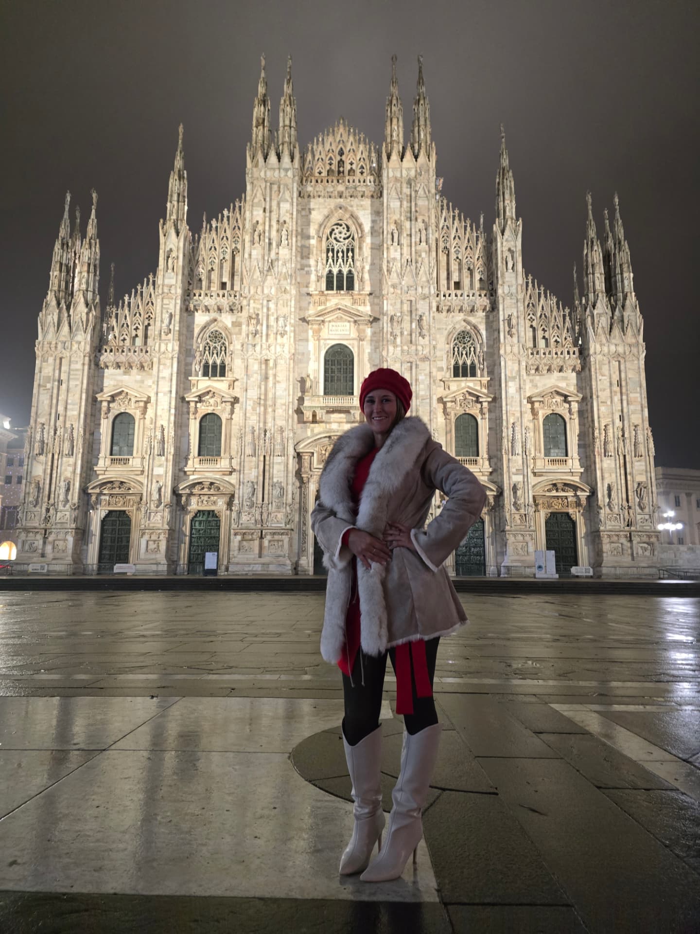 Nikki Bodie in a red coat standing on the empty piazza before the glowing Duomo di Milano late at night.