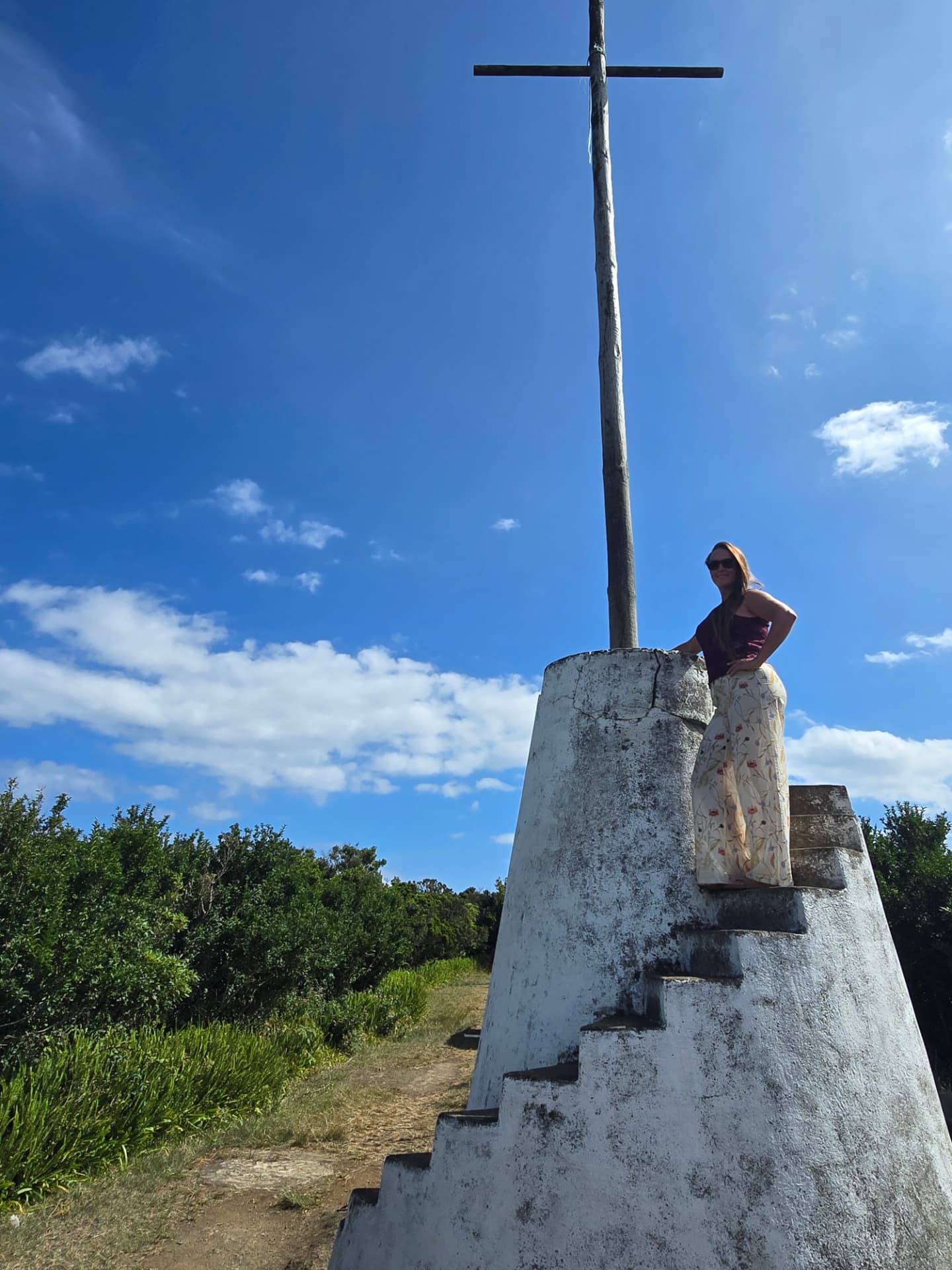 Nikki on the stepped white base of a tall wooden cross under a blue sky.