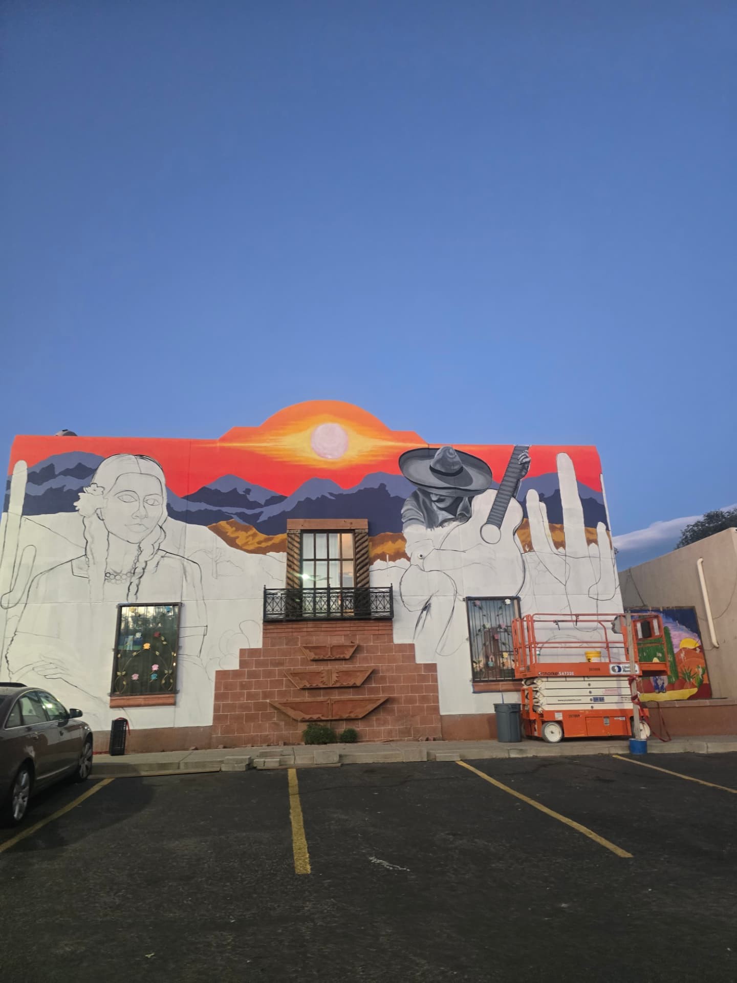 A half-finished mural of a desert sunset on a building wall at dusk.